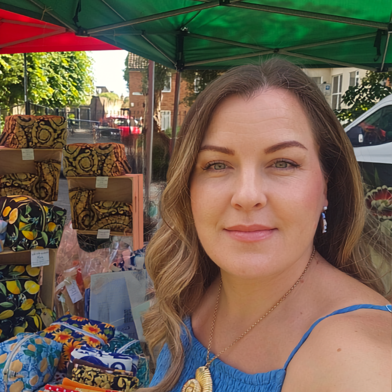 A woman with blonde-highlighted hair wearing a blue top and gold necklace, standing at an outdoor market stall under a green canopy with colourful patterned bags and clothing displayed behind her.