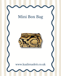 A mini box bag with a black and gold baroque scroll pattern, presented on a white card with a wavy blue border. The card features the text 'Mini Box Bag' at the top and 'www.